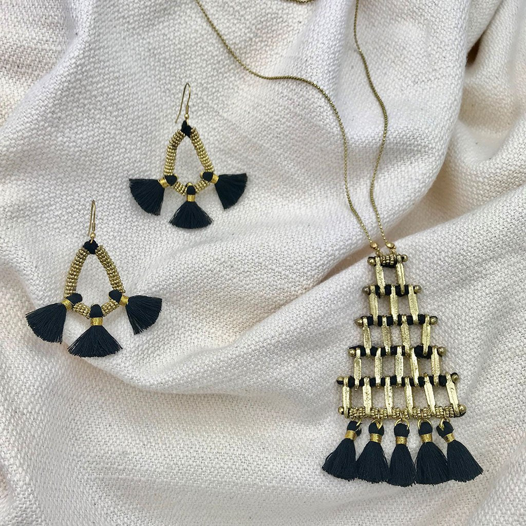 The Kai Pyramid Necklace is a gold-tone statement piece with a geometric grid design, featuring black tassels that gracefully hang at the bottom, adding an elegant touch.