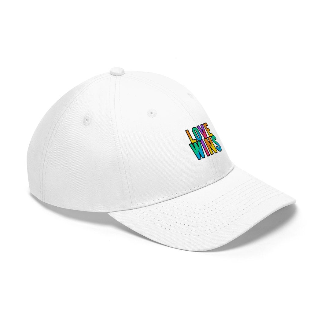 A bearded man wears the Rainbow Love Wins Unisex Twill Cap adorned with the phrase "Rainbow Love Wins" in vibrant colors, along with a gray sweatshirt, as he gazes into the distance against a simple backdrop.