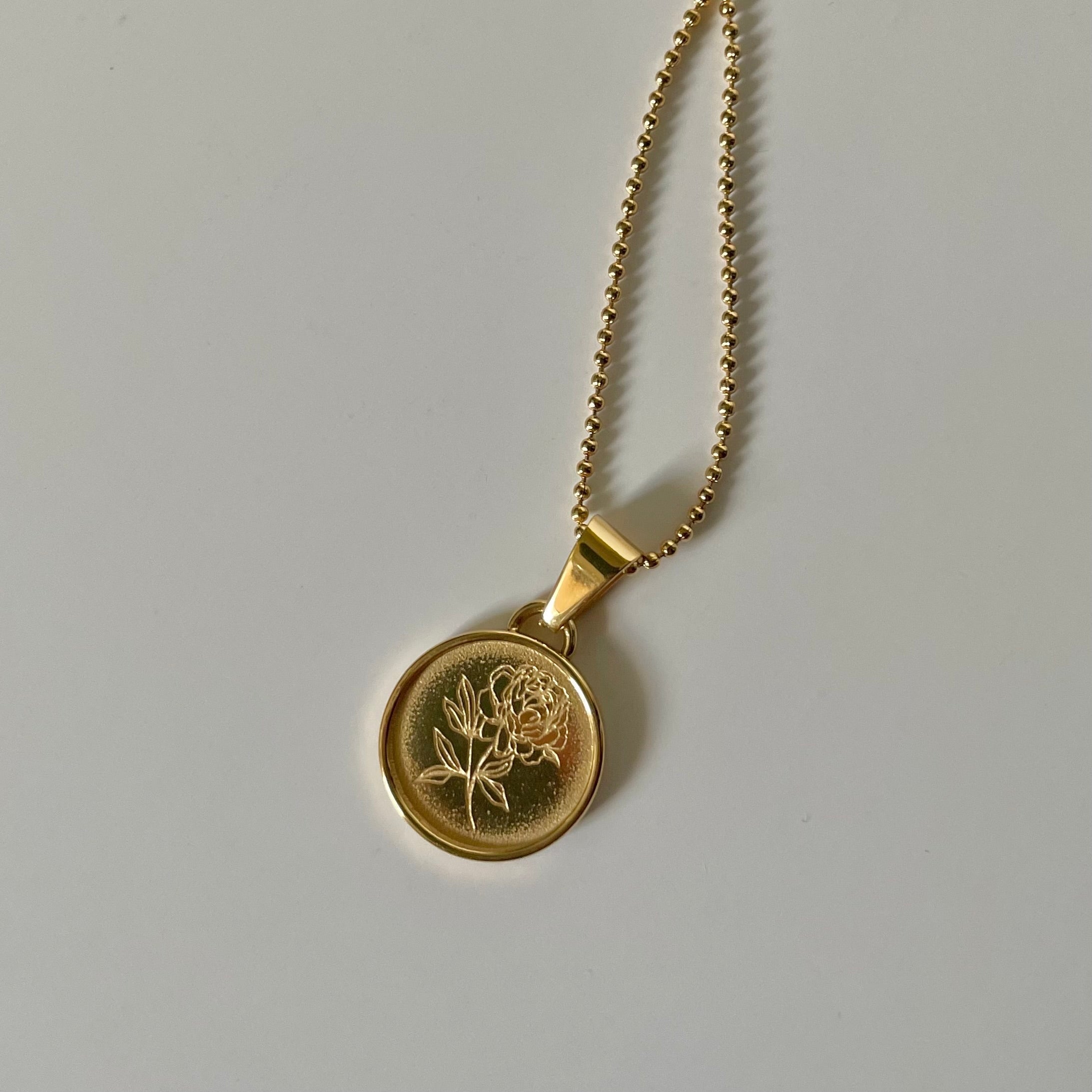 The Fleur De Pivione, also known as the Peony Necklace in Gold, features an 18K gold plated round pendant engraved with a delicate peony design, gracefully displayed on a white surface. This hypoallergenic jewelry piece provides both style and comfort for all-day wear.