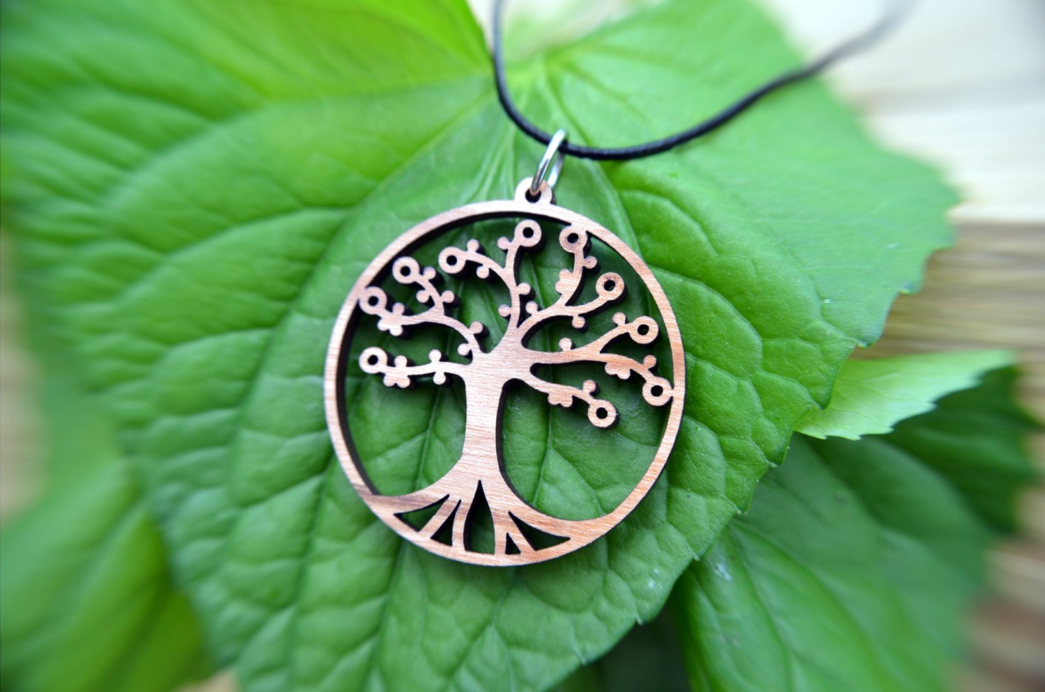 The Tree of Life Pendant in cherry, set on a leather cord and surrounded by lush green leaves, embodies the Natural Collection's commitment to organic designs.