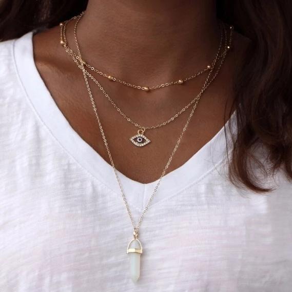 A person wearing the All Seeing Eye Multilayer Necklace with a gold eye-shaped pendant and another featuring a vertically elongated natural stone, possibly opal or amethyst, layered over a crisp white shirt.
