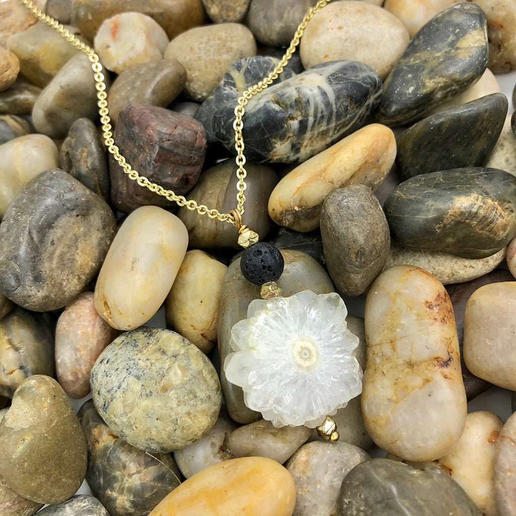 Introducing the Solar Quartz Lava Necklace: an artisan jewelry piece showcasing a gold chain adorned with a handmade white flower-shaped pendant and a small black bead.