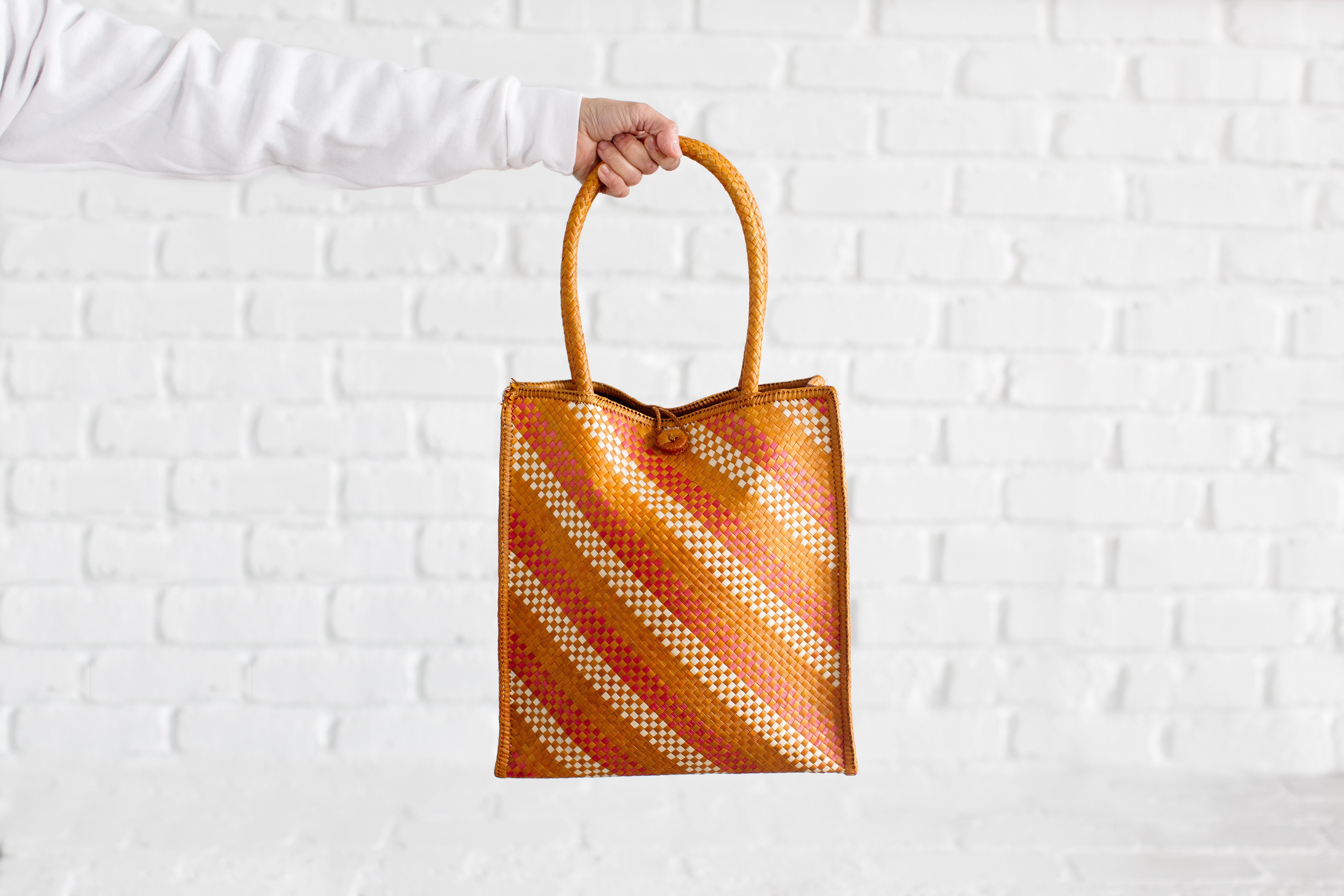 A hand holds the Handwoven Market Thip Tote, featuring orange, yellow, and white diagonal stripes against a white brick background. Expertly crafted by Chiangmai weavers, this tote seamlessly combines style and tradition.