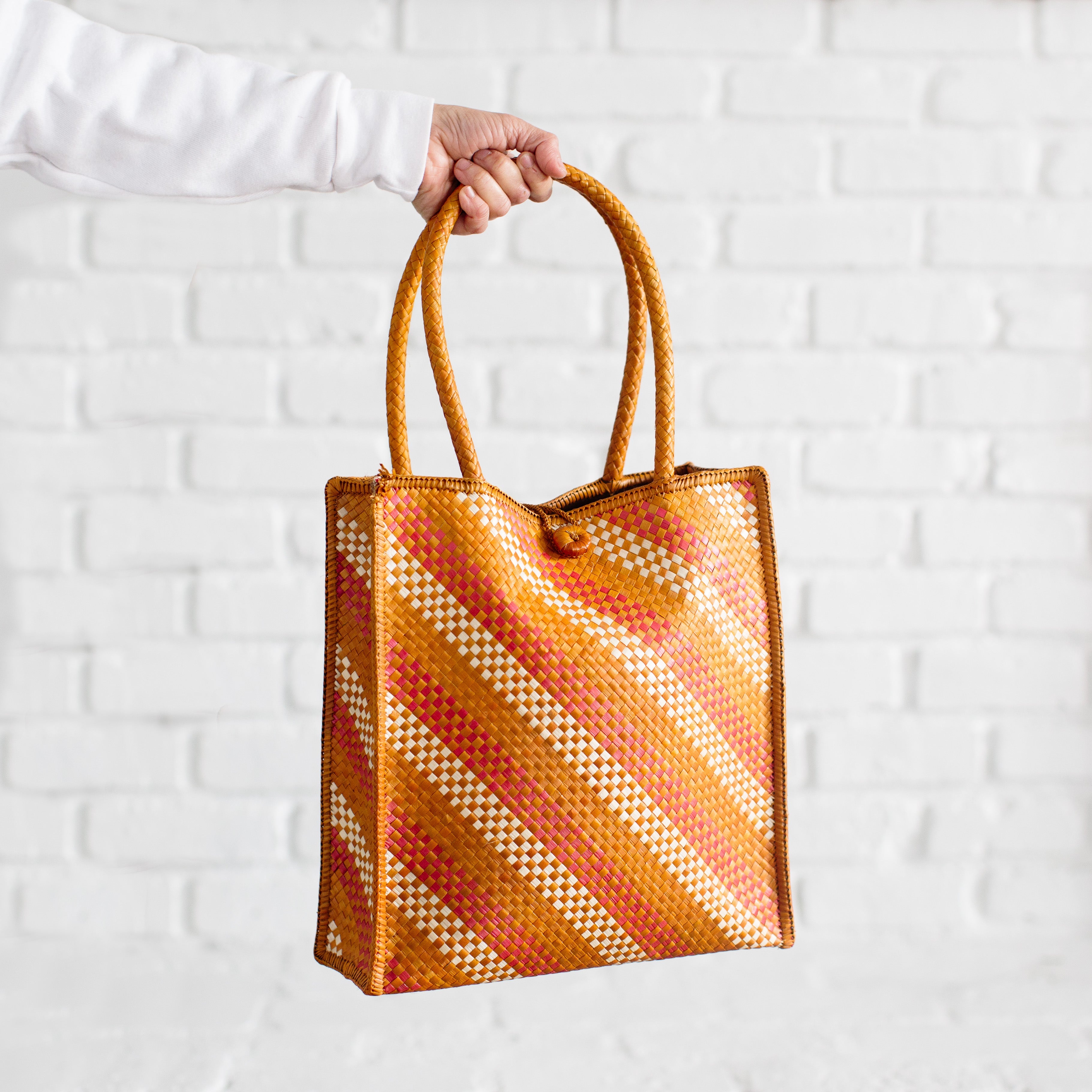 A hand holds the Handwoven Market Thip Tote, featuring orange, yellow, and white diagonal stripes against a white brick background. Expertly crafted by Chiangmai weavers, this tote seamlessly combines style and tradition.