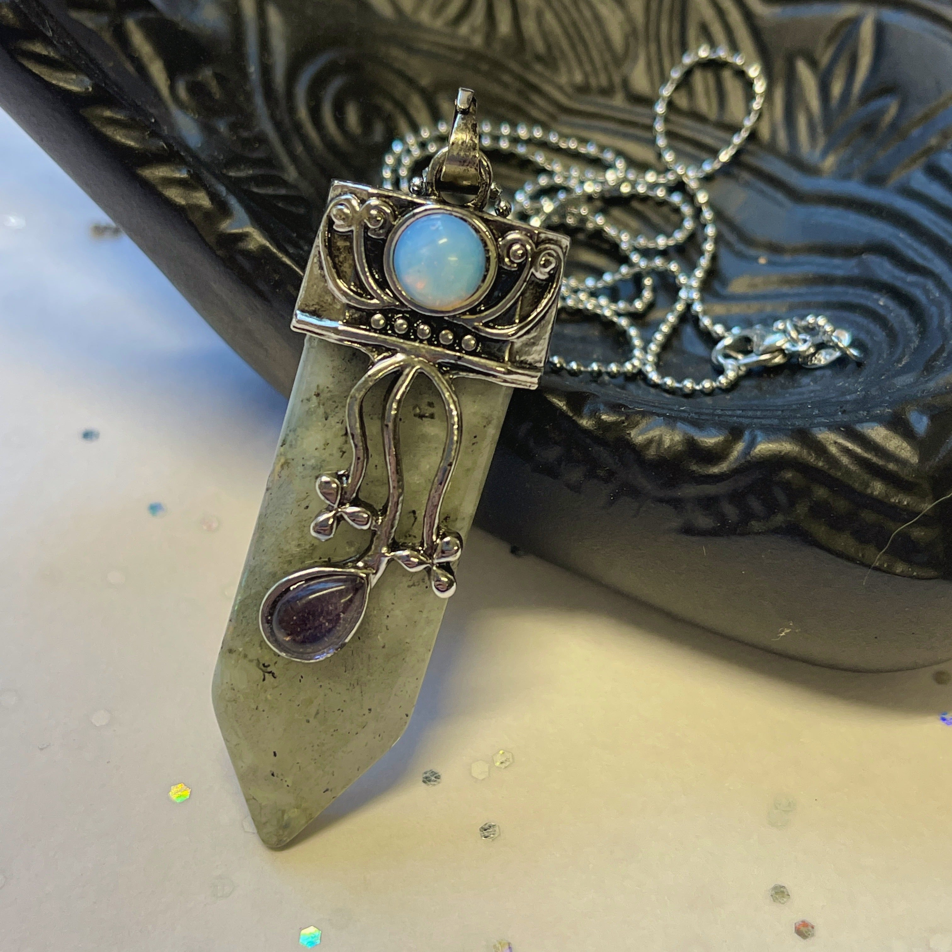 The Faerie Gemstone necklace features an enchanting opalite point adorned with intricate silver detailing, a small round moonstone, and a dangling amethyst. Accented with shimmering labradorite, it is elegantly showcased on a mannequin.