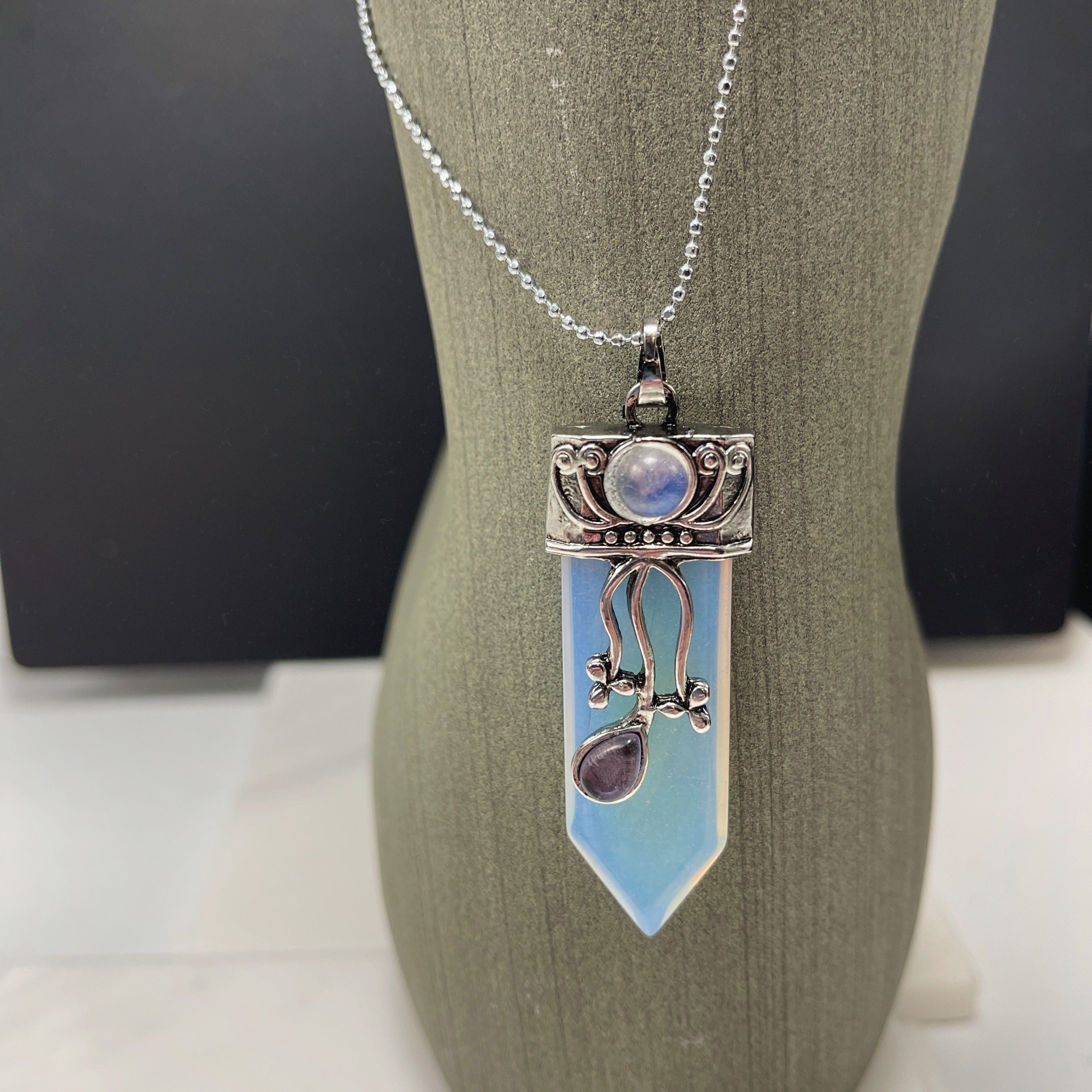 The Faerie Gemstone necklace features an enchanting opalite point adorned with intricate silver detailing, a small round moonstone, and a dangling amethyst. Accented with shimmering labradorite, it is elegantly showcased on a mannequin.