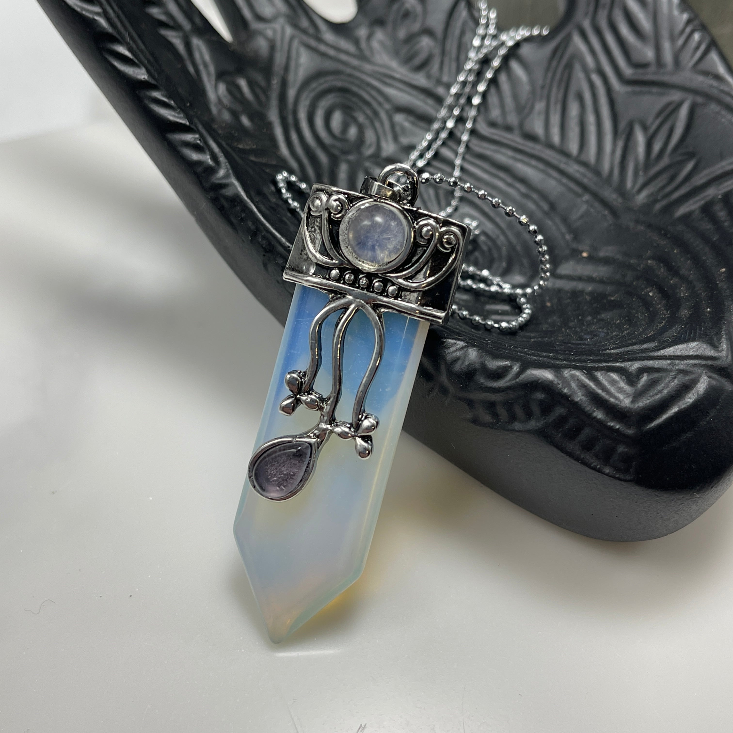 The Faerie Gemstone necklace features an enchanting opalite point adorned with intricate silver detailing, a small round moonstone, and a dangling amethyst. Accented with shimmering labradorite, it is elegantly showcased on a mannequin.