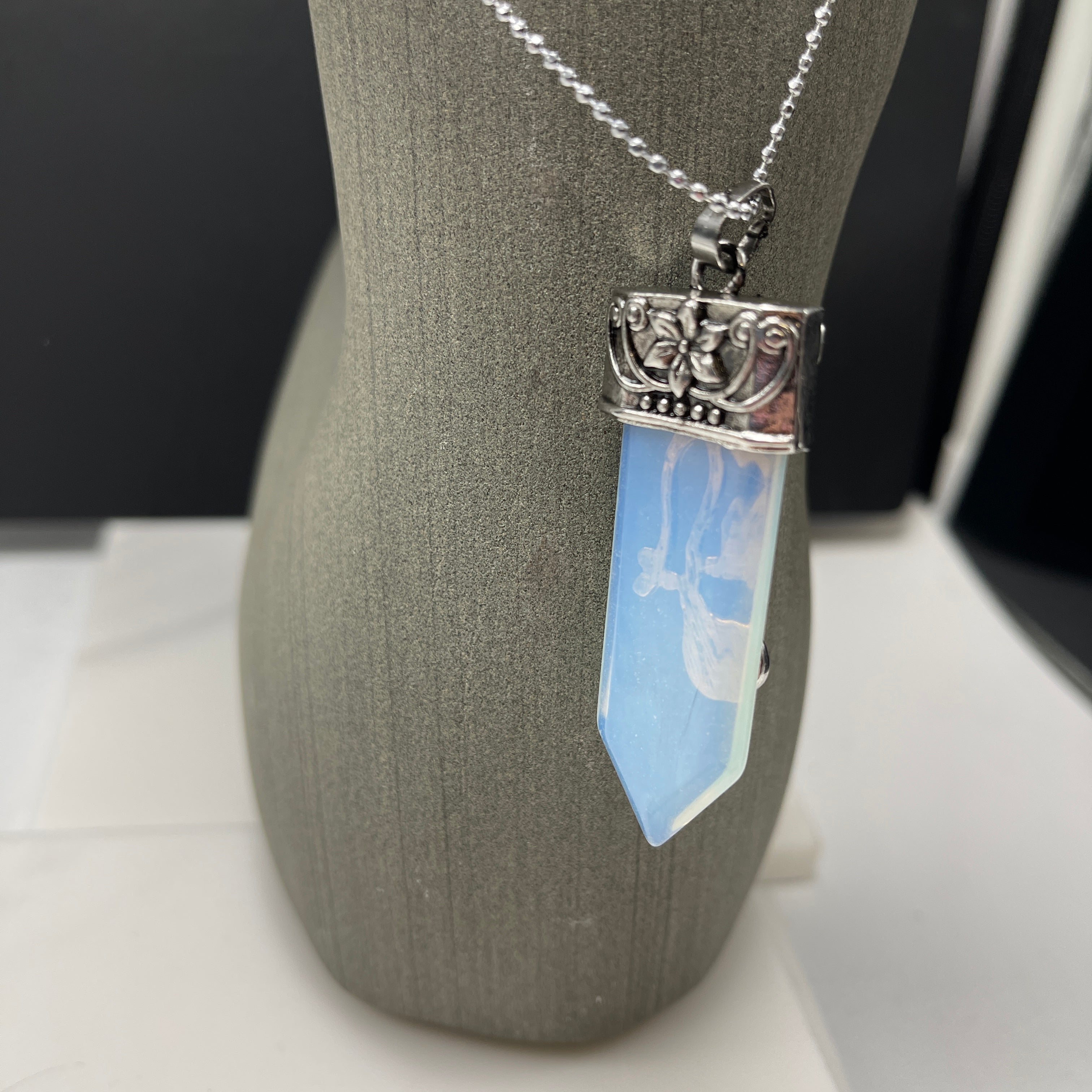 The Faerie Gemstone necklace features an enchanting opalite point adorned with intricate silver detailing, a small round moonstone, and a dangling amethyst. Accented with shimmering labradorite, it is elegantly showcased on a mannequin.