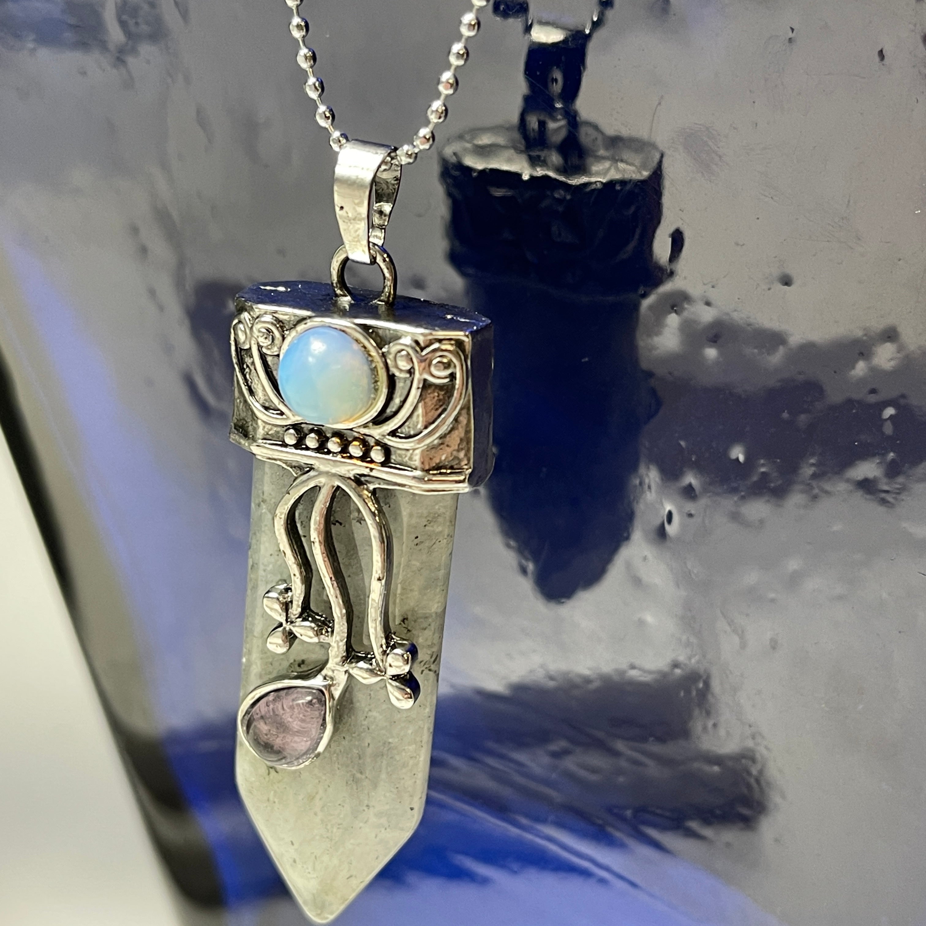 The Faerie Gemstone necklace features an enchanting opalite point adorned with intricate silver detailing, a small round moonstone, and a dangling amethyst. Accented with shimmering labradorite, it is elegantly showcased on a mannequin.