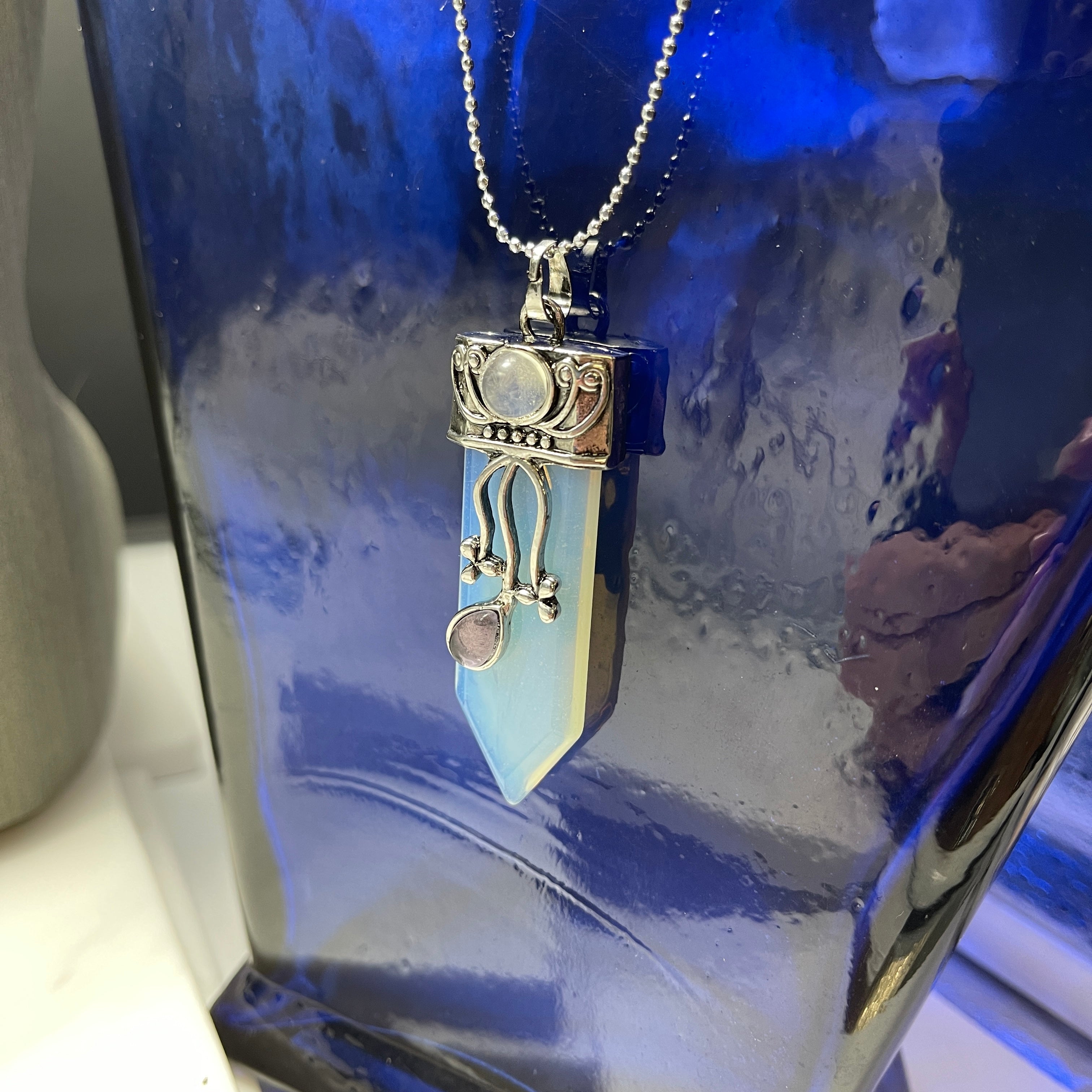 The Faerie Gemstone necklace features an enchanting opalite point adorned with intricate silver detailing, a small round moonstone, and a dangling amethyst. Accented with shimmering labradorite, it is elegantly showcased on a mannequin.