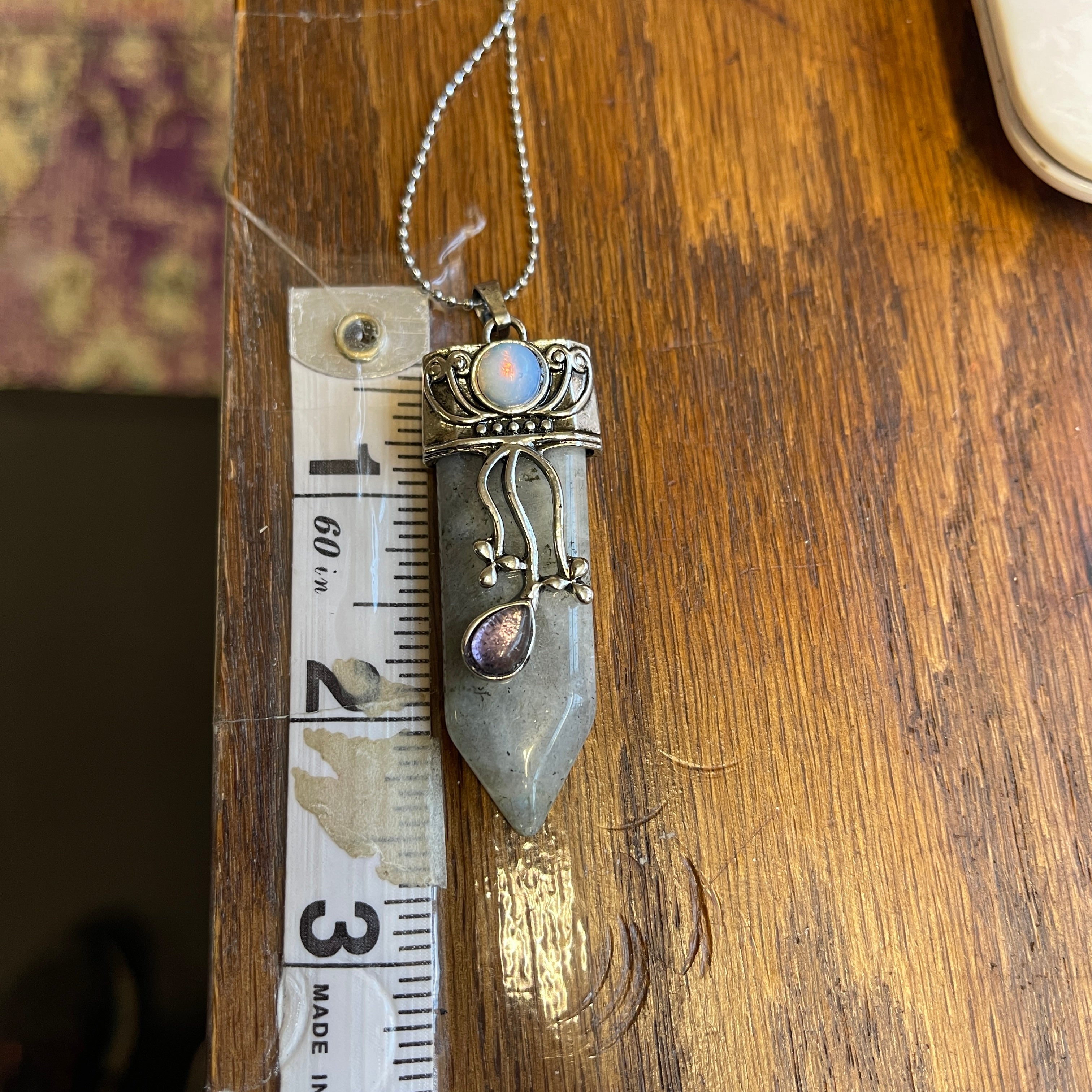 The Faerie Gemstone necklace features an enchanting opalite point adorned with intricate silver detailing, a small round moonstone, and a dangling amethyst. Accented with shimmering labradorite, it is elegantly showcased on a mannequin.