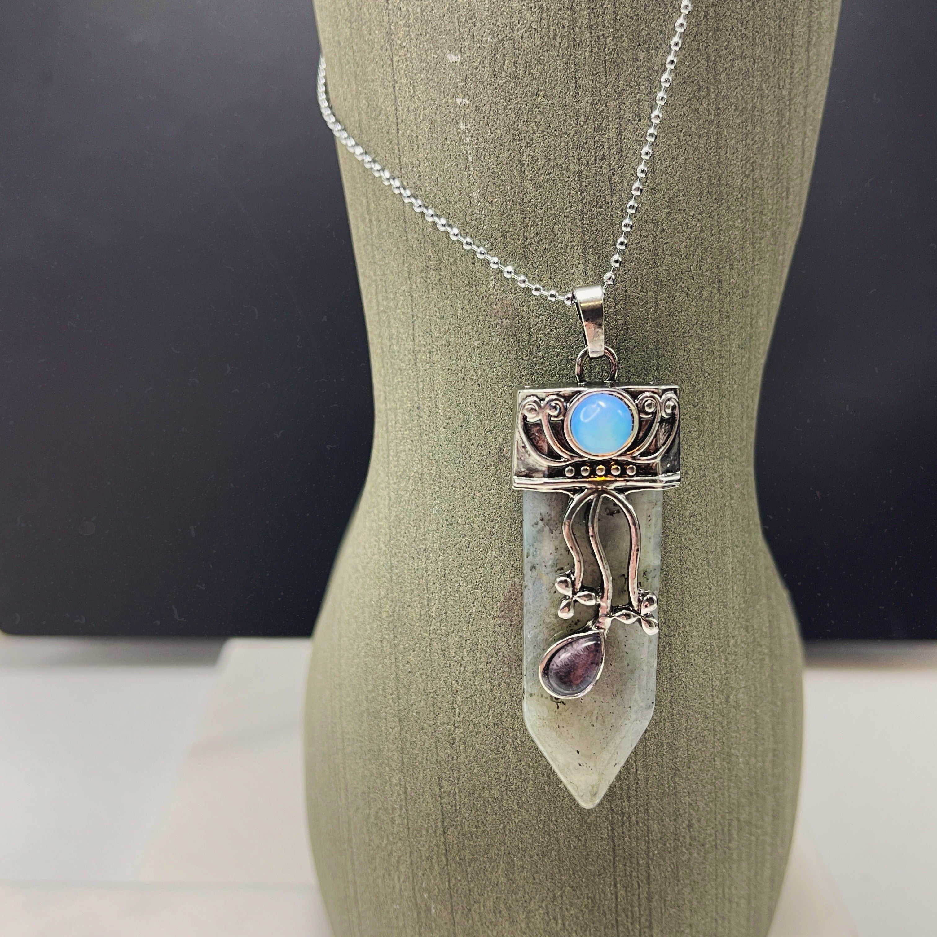 The Faerie Gemstone necklace features an enchanting opalite point adorned with intricate silver detailing, a small round moonstone, and a dangling amethyst. Accented with shimmering labradorite, it is elegantly showcased on a mannequin.