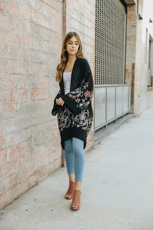 A woman in a Long Floral Kimono Cardigan stands by a brick wall, glancing over her shoulder.