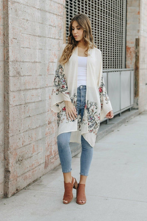 A woman in a Long Floral Kimono Cardigan stands by a brick wall, glancing over her shoulder.