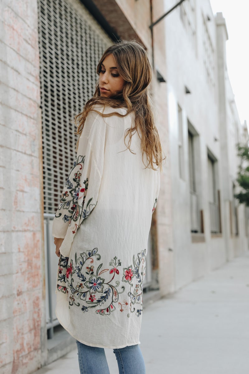 A woman in a Long Floral Kimono Cardigan stands by a brick wall, glancing over her shoulder.