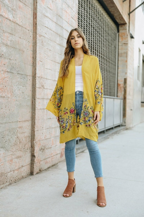 A woman in a Long Floral Kimono Cardigan stands by a brick wall, glancing over her shoulder.