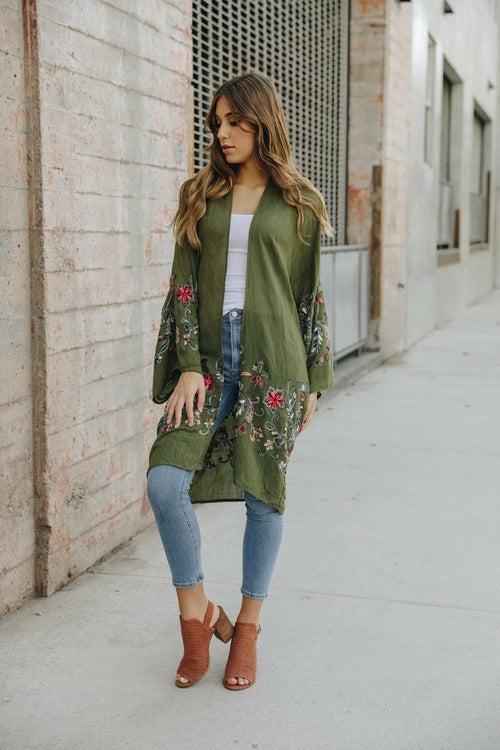 A woman in a Long Floral Kimono Cardigan stands by a brick wall, glancing over her shoulder.