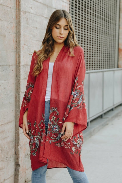 A woman in a Long Floral Kimono Cardigan stands by a brick wall, glancing over her shoulder.