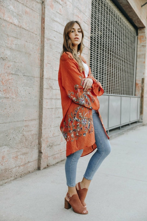 A woman in a Long Floral Kimono Cardigan stands by a brick wall, glancing over her shoulder.