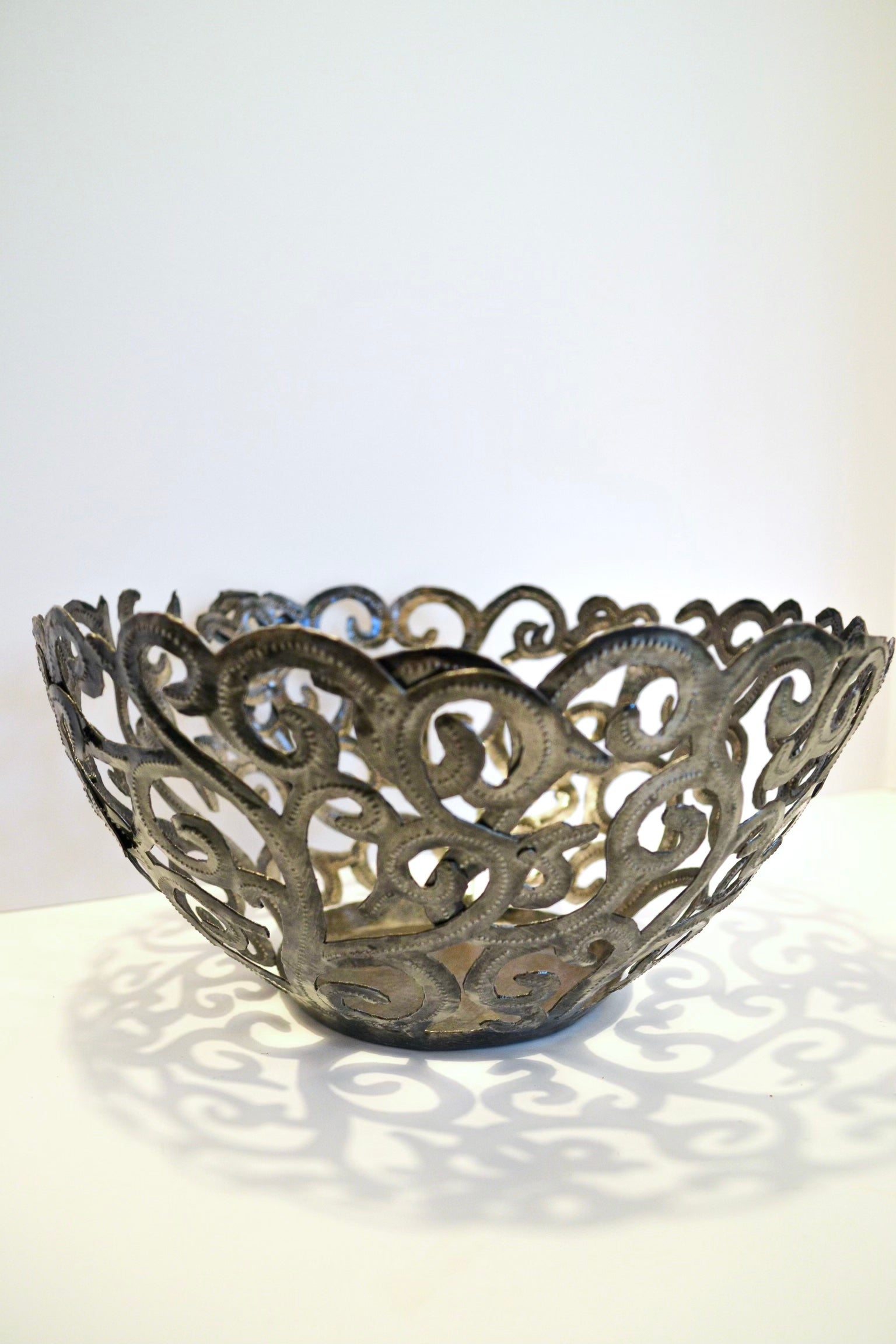 The Metal Art Scroll Bowl, intricately designed with swirling cut-out patterns and crafted from recycled steel, is elegantly displayed on a white surface.
