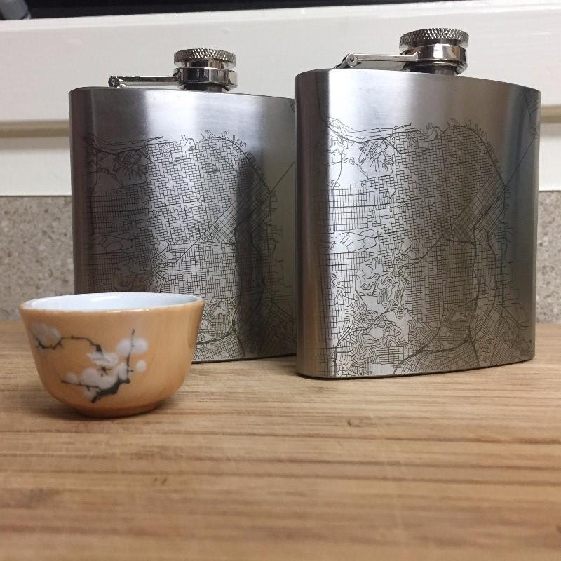 The Parker - Colorado Map Hip Flask lies on a wooden surface, accompanied by a small metal funnel and a white cloth drawstring bag.