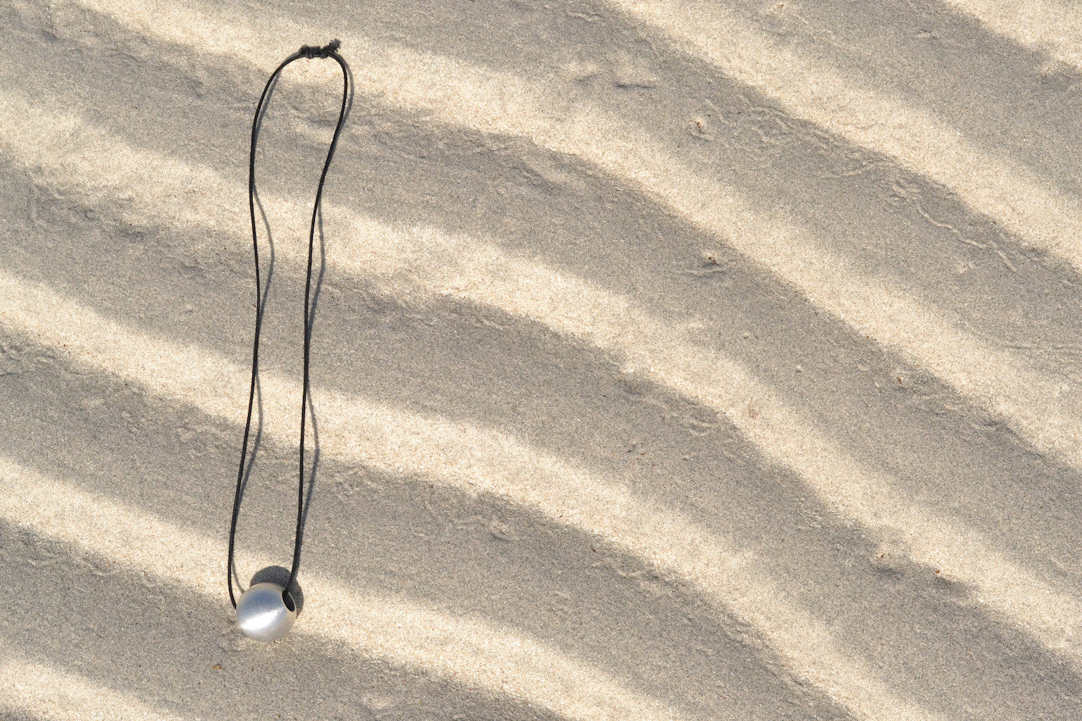 The Recycled Bombshell Ball Necklace is a stunning spherical pendant made from silver, hanging on a black cord. It is meticulously handcrafted by artisans in Laos using recycled bomb materials. Displayed against a white background, the necklace elegantly highlights the unique beauty of artisan jewelry.