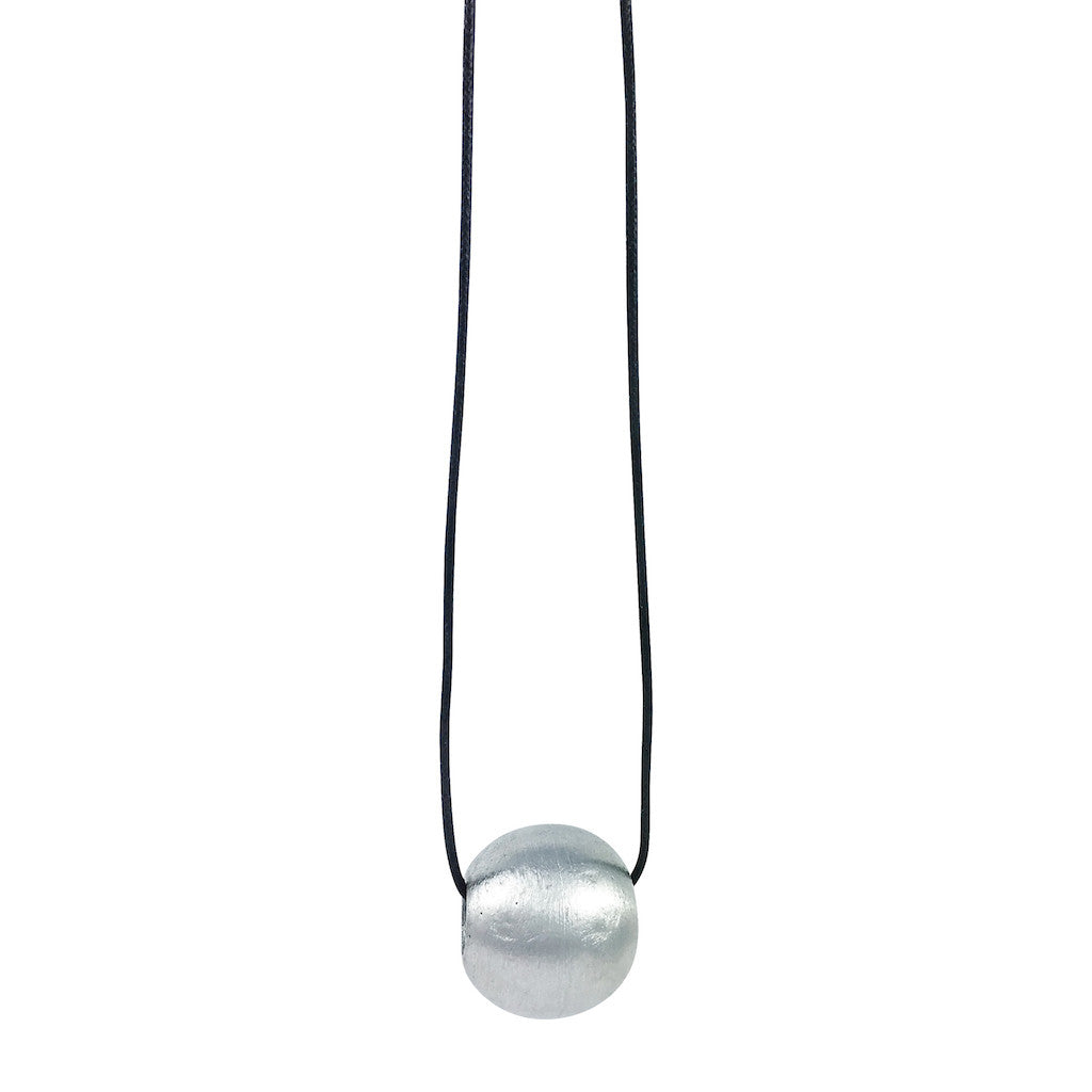The Recycled Bombshell Ball Necklace is a stunning spherical pendant made from silver, hanging on a black cord. It is meticulously handcrafted by artisans in Laos using recycled bomb materials. Displayed against a white background, the necklace elegantly highlights the unique beauty of artisan jewelry.