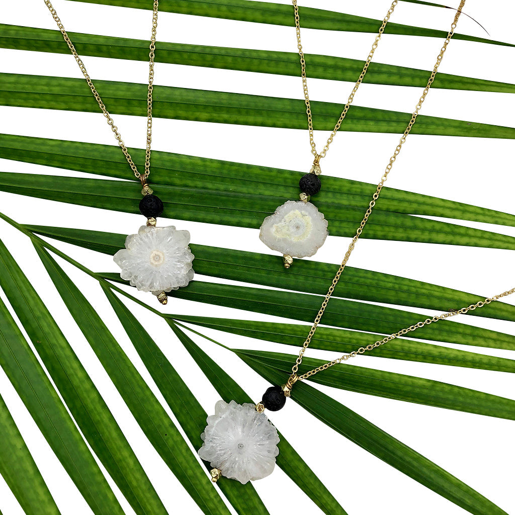 Introducing the Solar Quartz Lava Necklace: an artisan jewelry piece showcasing a gold chain adorned with a handmade white flower-shaped pendant and a small black bead.