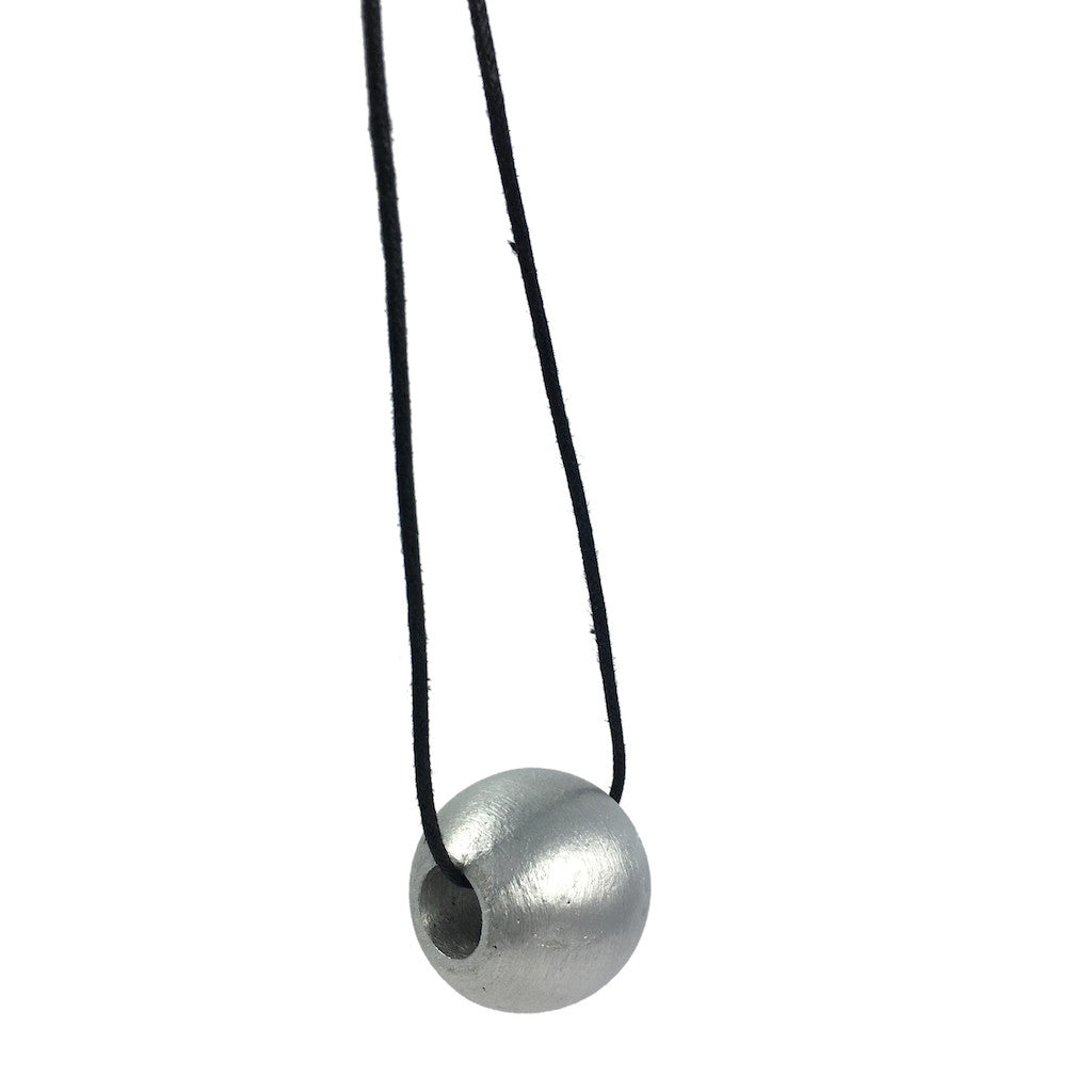 The Recycled Bombshell Ball Necklace is a stunning spherical pendant made from silver, hanging on a black cord. It is meticulously handcrafted by artisans in Laos using recycled bomb materials. Displayed against a white background, the necklace elegantly highlights the unique beauty of artisan jewelry.