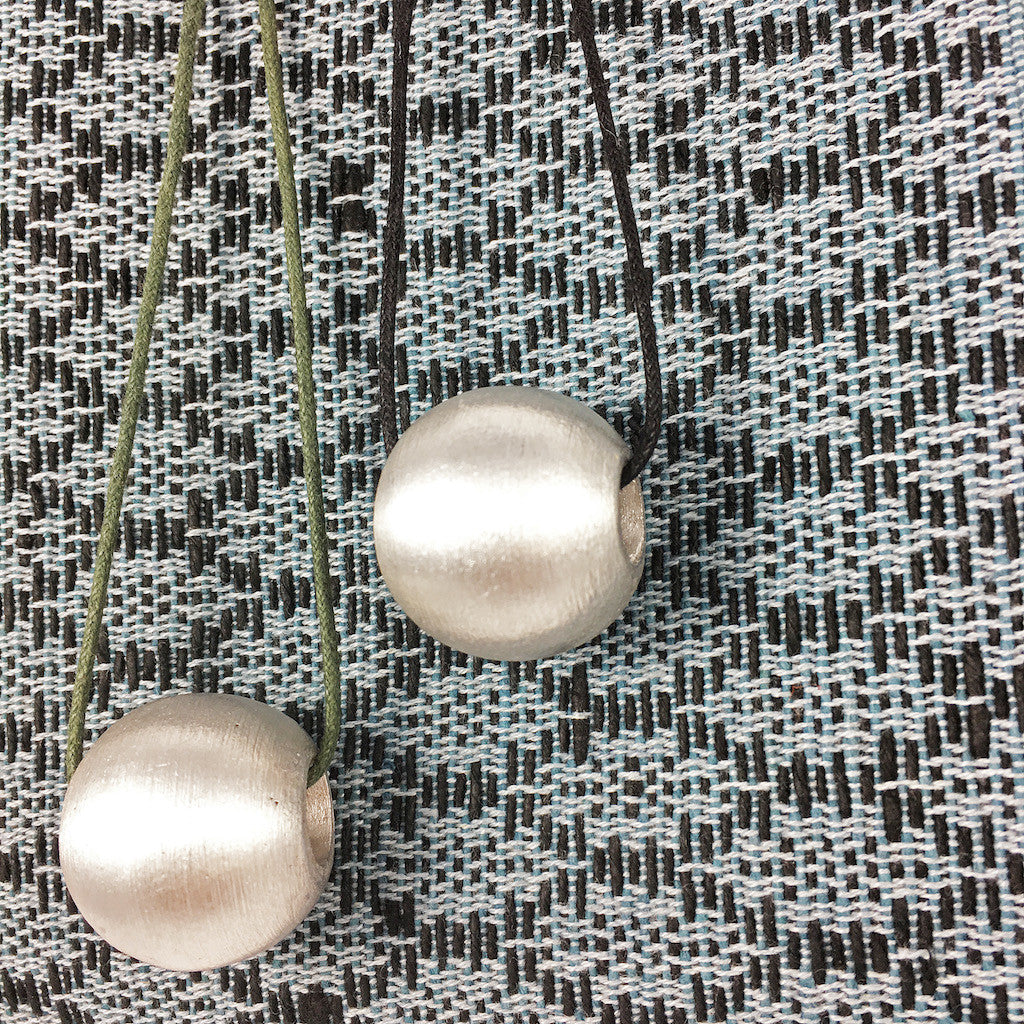 The Recycled Bombshell Ball Necklace is a stunning spherical pendant made from silver, hanging on a black cord. It is meticulously handcrafted by artisans in Laos using recycled bomb materials. Displayed against a white background, the necklace elegantly highlights the unique beauty of artisan jewelry.