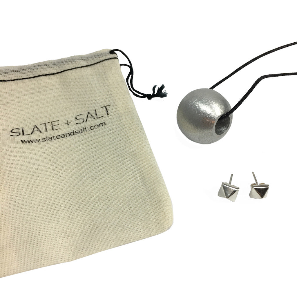 The Recycled Bombshell Ball Necklace is a stunning spherical pendant made from silver, hanging on a black cord. It is meticulously handcrafted by artisans in Laos using recycled bomb materials. Displayed against a white background, the necklace elegantly highlights the unique beauty of artisan jewelry.