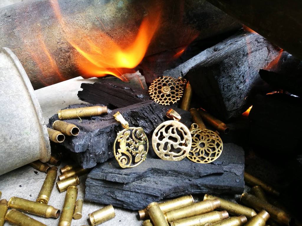 Crafted in Cambodia from recycled brass bombshells, the Bombshell Peace Earrings feature a gold-colored circular peace sign design with cutouts and hook attachments.