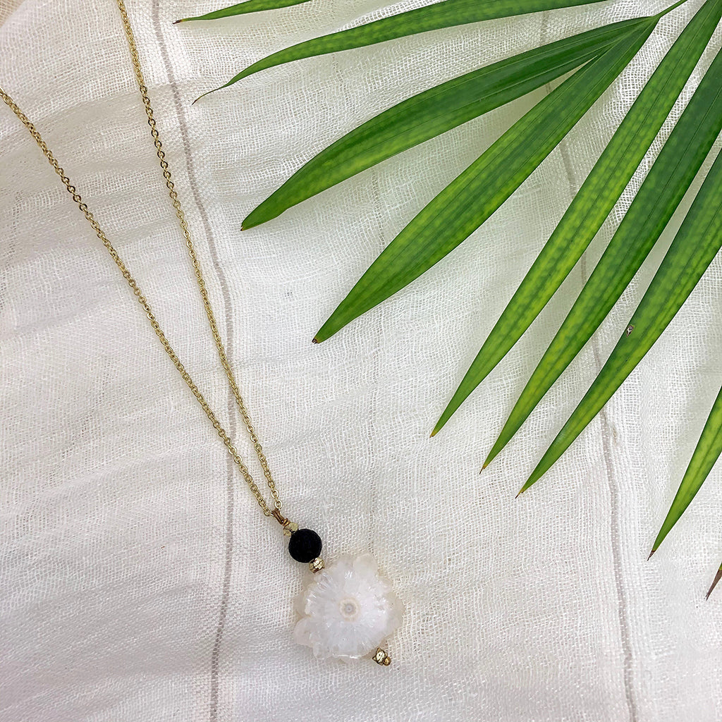 Introducing the Solar Quartz Lava Necklace: an artisan jewelry piece showcasing a gold chain adorned with a handmade white flower-shaped pendant and a small black bead.