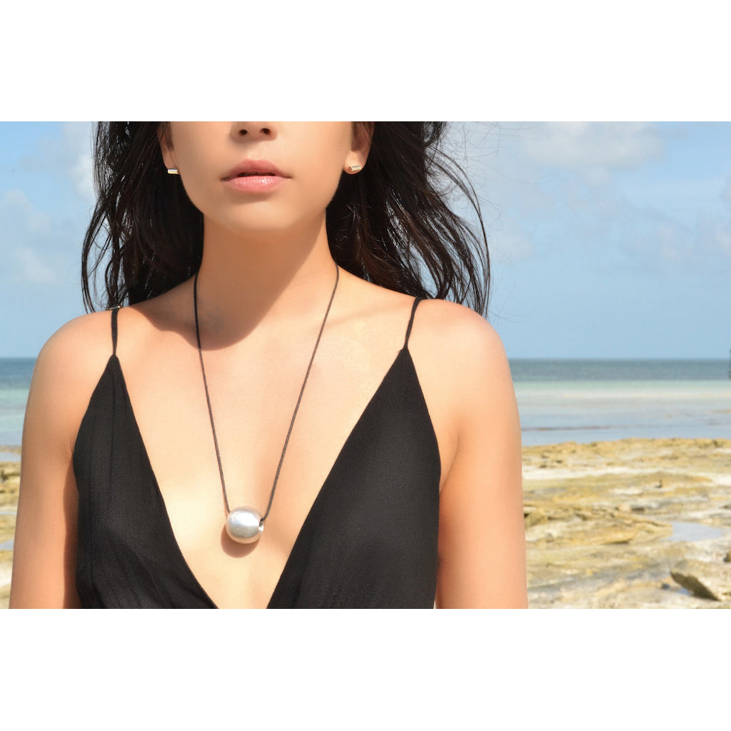 The Recycled Bombshell Ball Necklace is a stunning spherical pendant made from silver, hanging on a black cord. It is meticulously handcrafted by artisans in Laos using recycled bomb materials. Displayed against a white background, the necklace elegantly highlights the unique beauty of artisan jewelry.