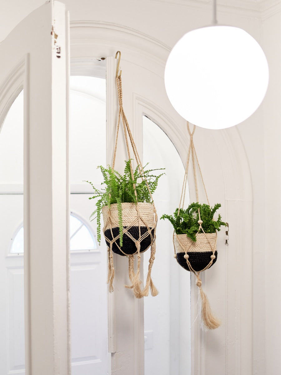 The Plant Hanger - Nadu features a woven basket with a black and natural fiber design, suspended by a handmade jute hanger adorned with braided and knotted ropes, complete with a tassel at the bottom.