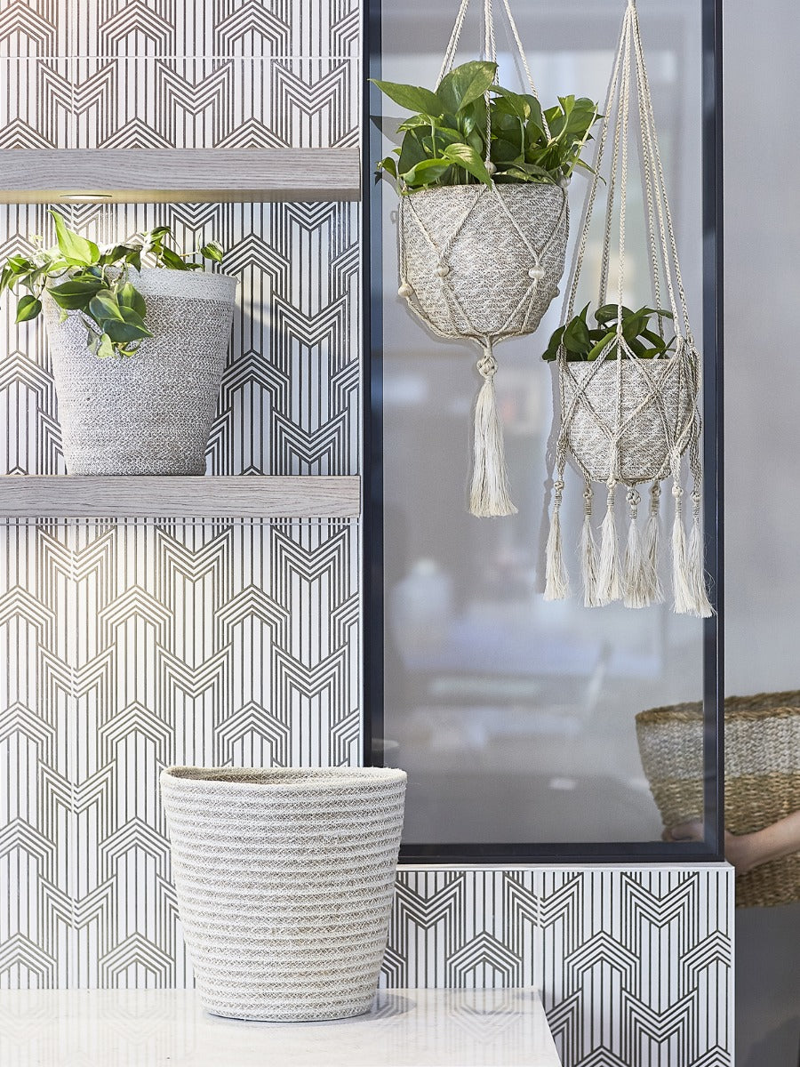 The Plant Hanger - Nadu features a woven basket with a black and natural fiber design, suspended by a handmade jute hanger adorned with braided and knotted ropes, complete with a tassel at the bottom.