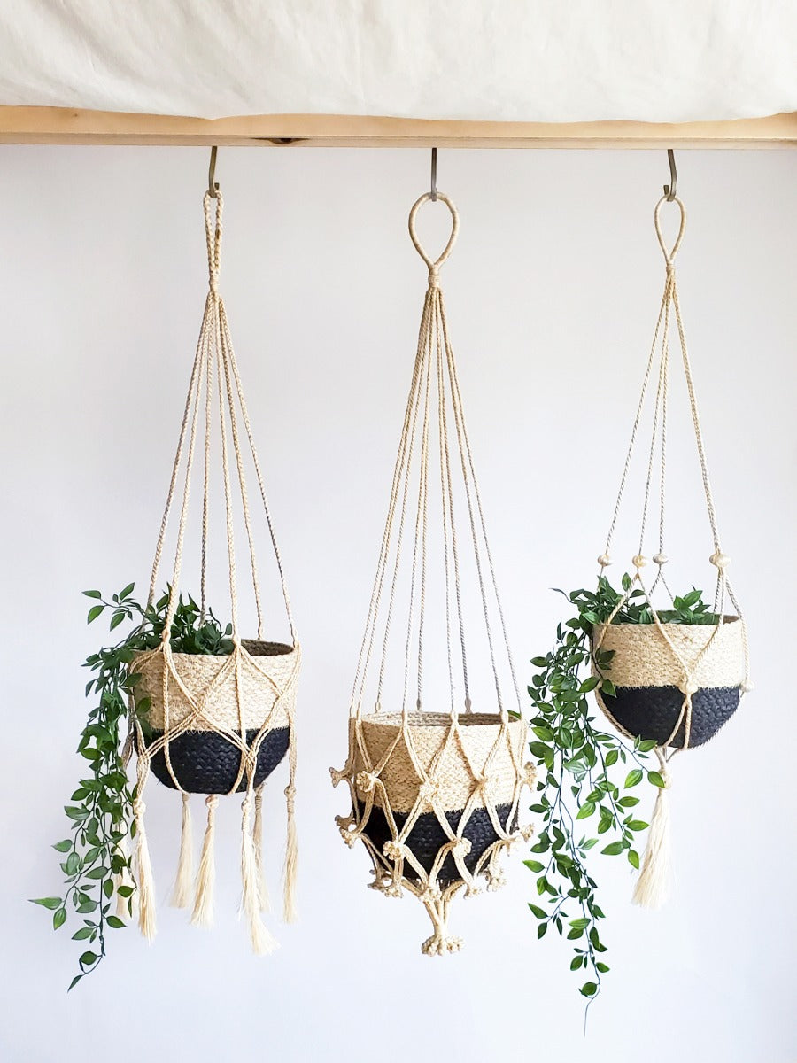 The Plant Hanger - Nadu features a woven basket with a black and natural fiber design, suspended by a handmade jute hanger adorned with braided and knotted ropes, complete with a tassel at the bottom.