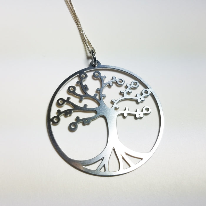 The Tree of Life Pendant in cherry, set on a leather cord and surrounded by lush green leaves, embodies the Natural Collection's commitment to organic designs.