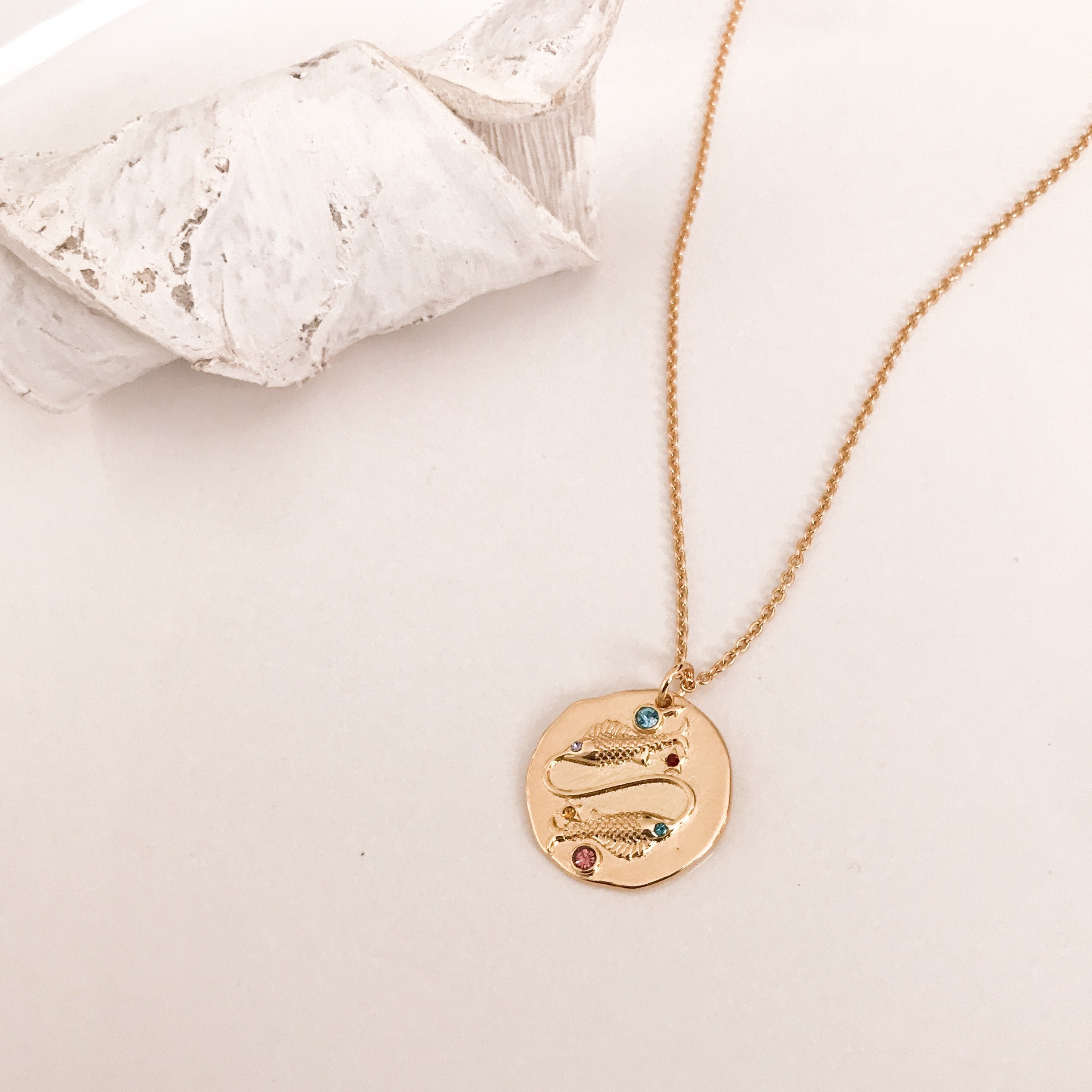 Discover our Wanderlust Zodiac Necklace collection, featuring exquisite hanging gold pendants with intricate designs on delicate chains. Some necklaces are embellished with vibrant gemstones. Crafted with hypoallergenic 18K gold plating, these pieces effortlessly combine elegance and comfort for everyday wear.