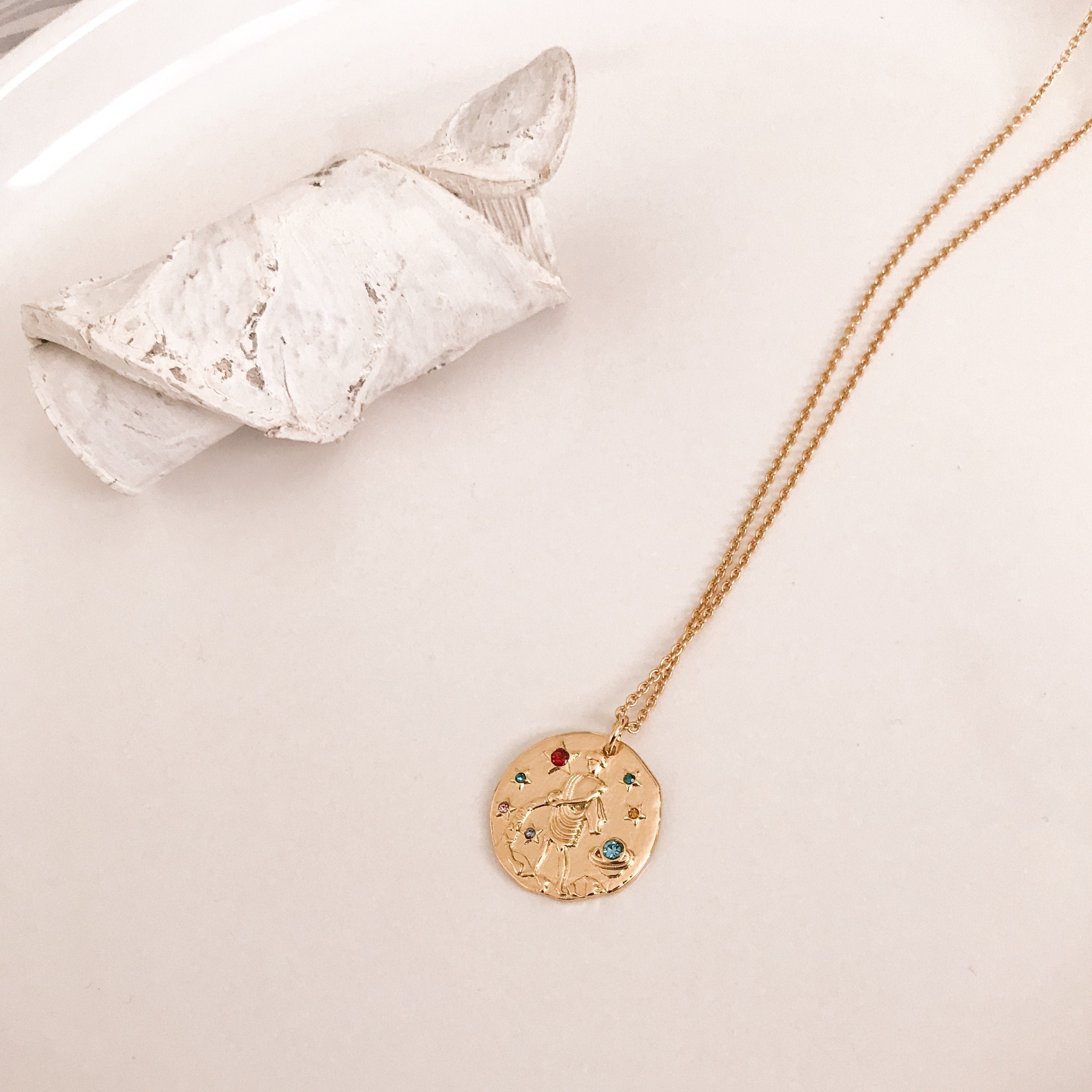Discover our Wanderlust Zodiac Necklace collection, featuring exquisite hanging gold pendants with intricate designs on delicate chains. Some necklaces are embellished with vibrant gemstones. Crafted with hypoallergenic 18K gold plating, these pieces effortlessly combine elegance and comfort for everyday wear.