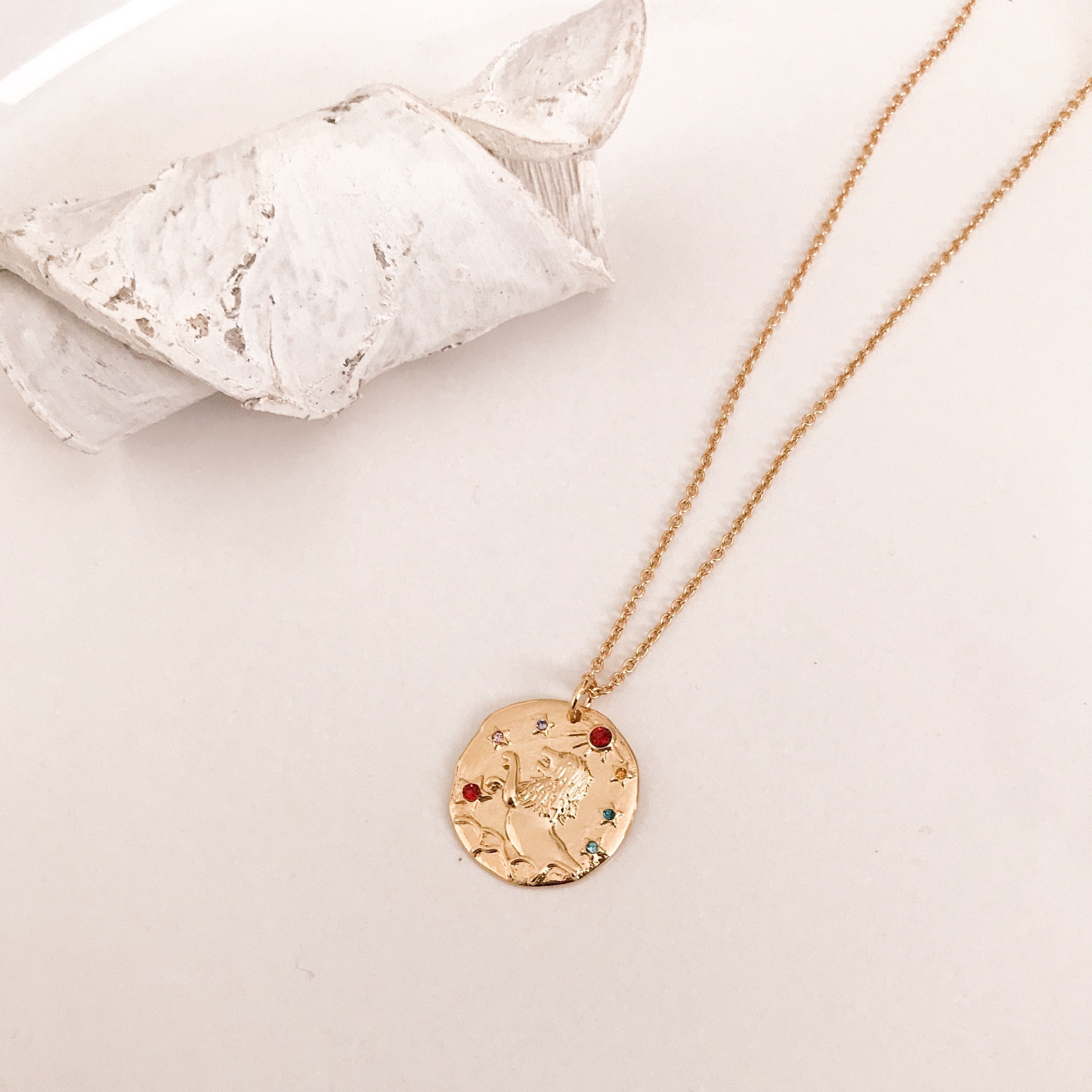 Discover our Wanderlust Zodiac Necklace collection, featuring exquisite hanging gold pendants with intricate designs on delicate chains. Some necklaces are embellished with vibrant gemstones. Crafted with hypoallergenic 18K gold plating, these pieces effortlessly combine elegance and comfort for everyday wear.
