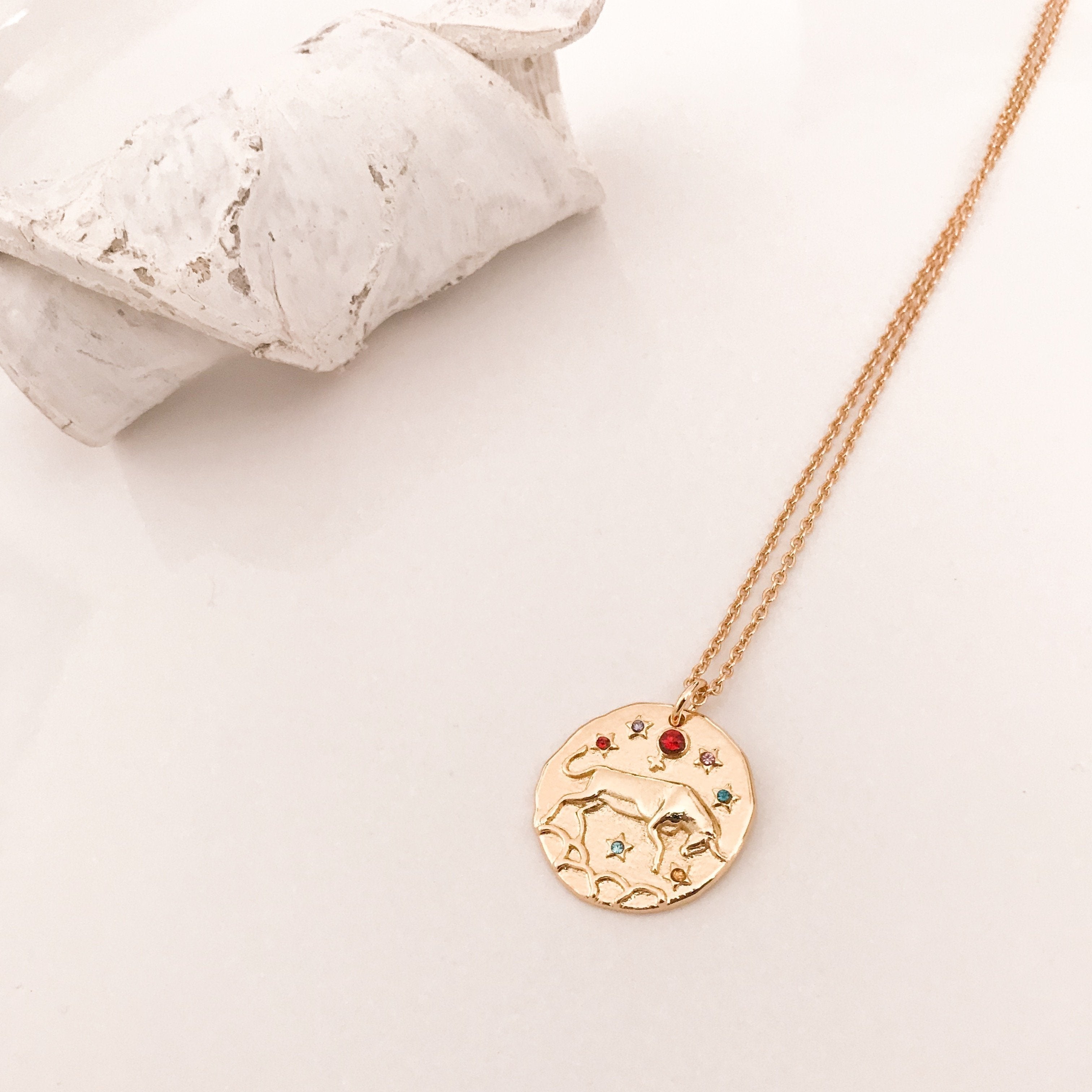 Discover our Wanderlust Zodiac Necklace collection, featuring exquisite hanging gold pendants with intricate designs on delicate chains. Some necklaces are embellished with vibrant gemstones. Crafted with hypoallergenic 18K gold plating, these pieces effortlessly combine elegance and comfort for everyday wear.
