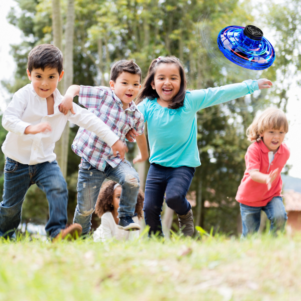 Children playing tag in a lush green park, with one girl in blue shorts reaching to tag a boy in a striped shirt, while a Glow Drone 360 Flying Toy with LED lights hovers above.