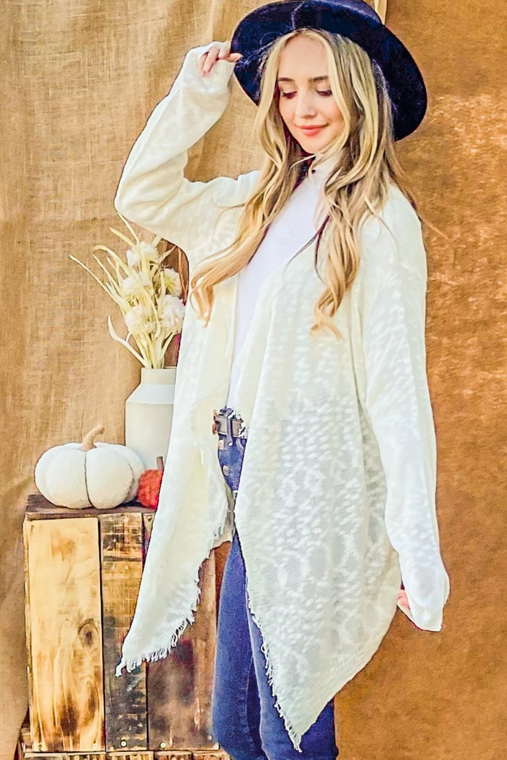 A person with long blonde hair wears a black hat, the And The Why Textured Knit Fringe Hem Cardigan in white, and blue jeans, standing against a brown backdrop near a wooden table with a vase of dried flowers and a small pumpkin.