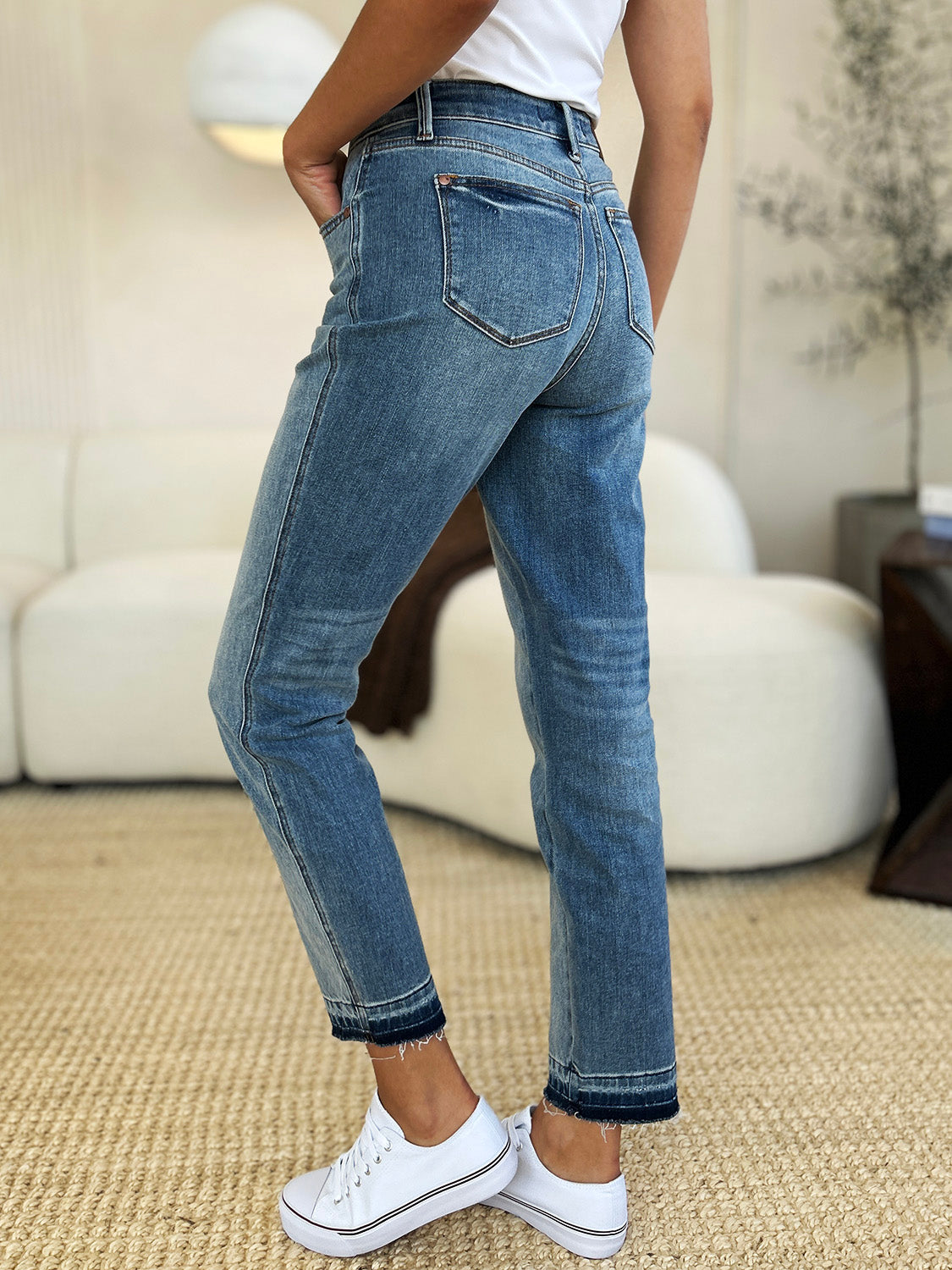 Person wearing a black top, Judy Blue Full Size Mid Rise Rigid Magic Release Hem Jeans, and black sneakers with white laces, standing on a beige carpet with hands in pockets.