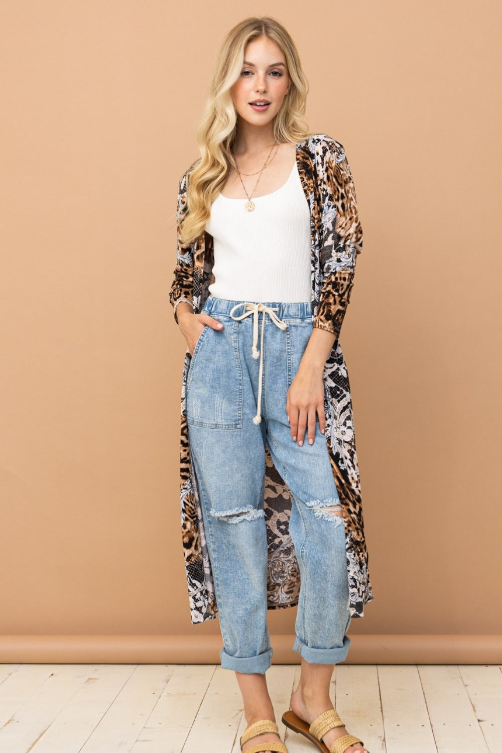 A woman with blonde hair wears a white tank top, the And The Why Leopard Kimono Open Front Longline Cardigan, light blue drawstring jeans, and sandals, standing against a beige background.