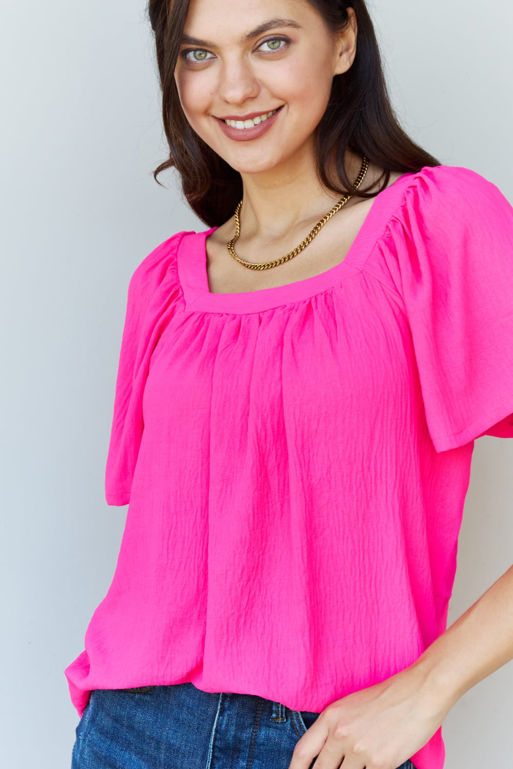 A person wearing the Ninexis Keep Me Close Square Neck Short Sleeve Blouse in Fuchsia paired with blue jeans smiles while posing against a plain background.