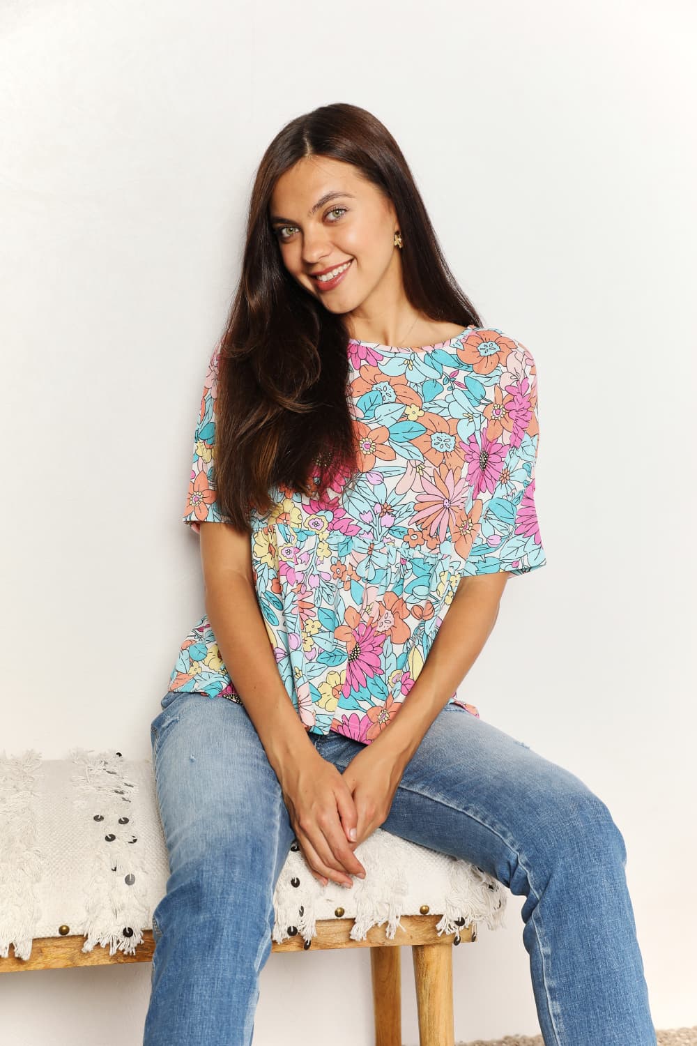 A person wearing a Double Take Floral Round Neck Babydoll Top and jeans stands with one hand in their pocket, with a plant in the background.