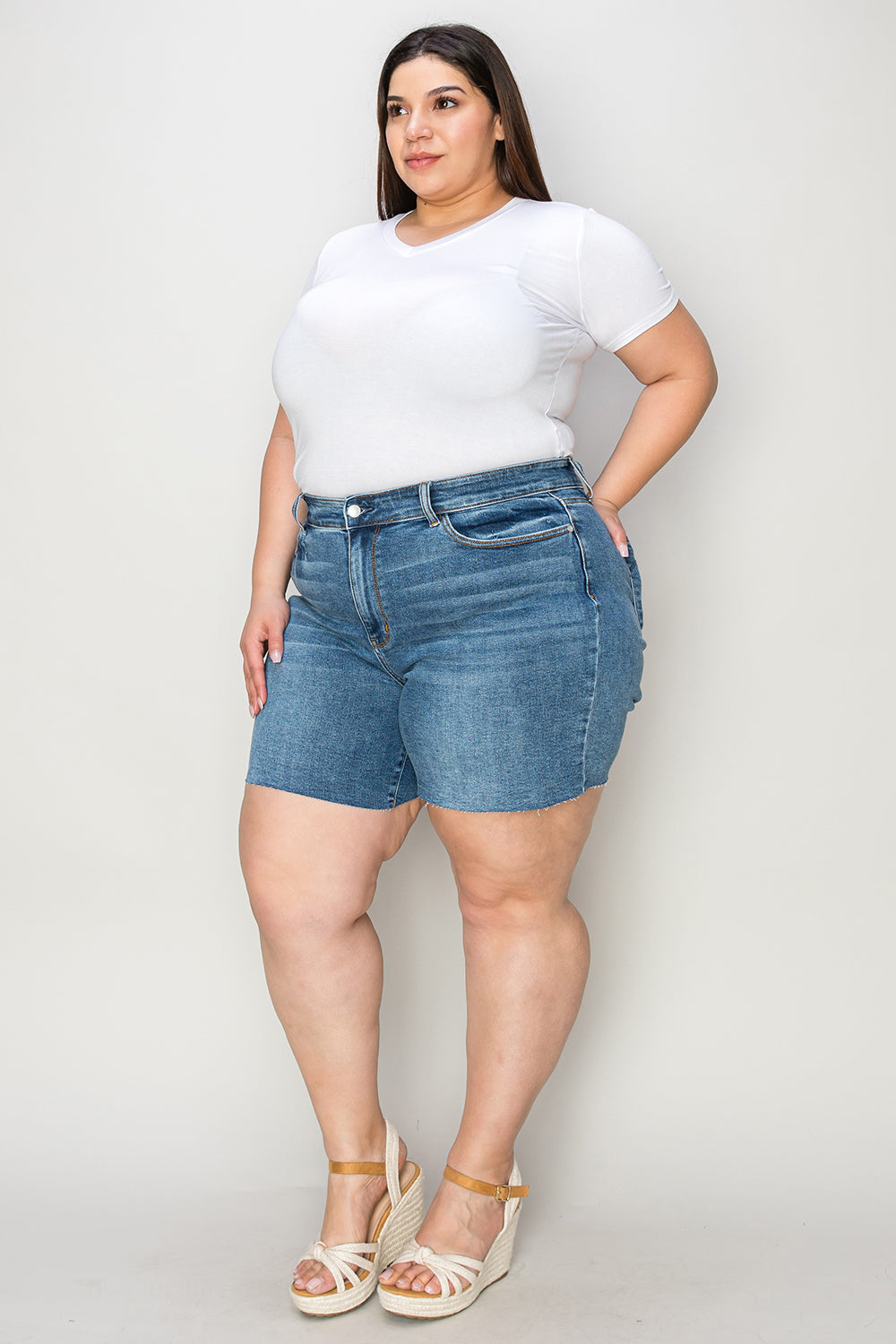 A woman in a white short-sleeve top and Judy Blue Full Size High Waist Slim Denim Shorts stands against a plain backdrop. She is smiling with her hands resting on her hips, wearing white open-toe wedge sandals that emphasize the quality denim fabric of her high-waisted design.