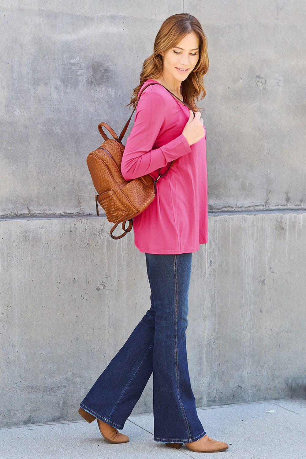 A woman with light brown hair stands against a concrete wall, wearing the Basic Bae Full Size V-Neck Long Sleeve Top in olive green and blue jeans, with a white backpack on her shoulder. Her outfit exudes basic style and is made from polyester, ensuring it’s both comfortable and suitable for machine wash cold. She is looking at the camera.